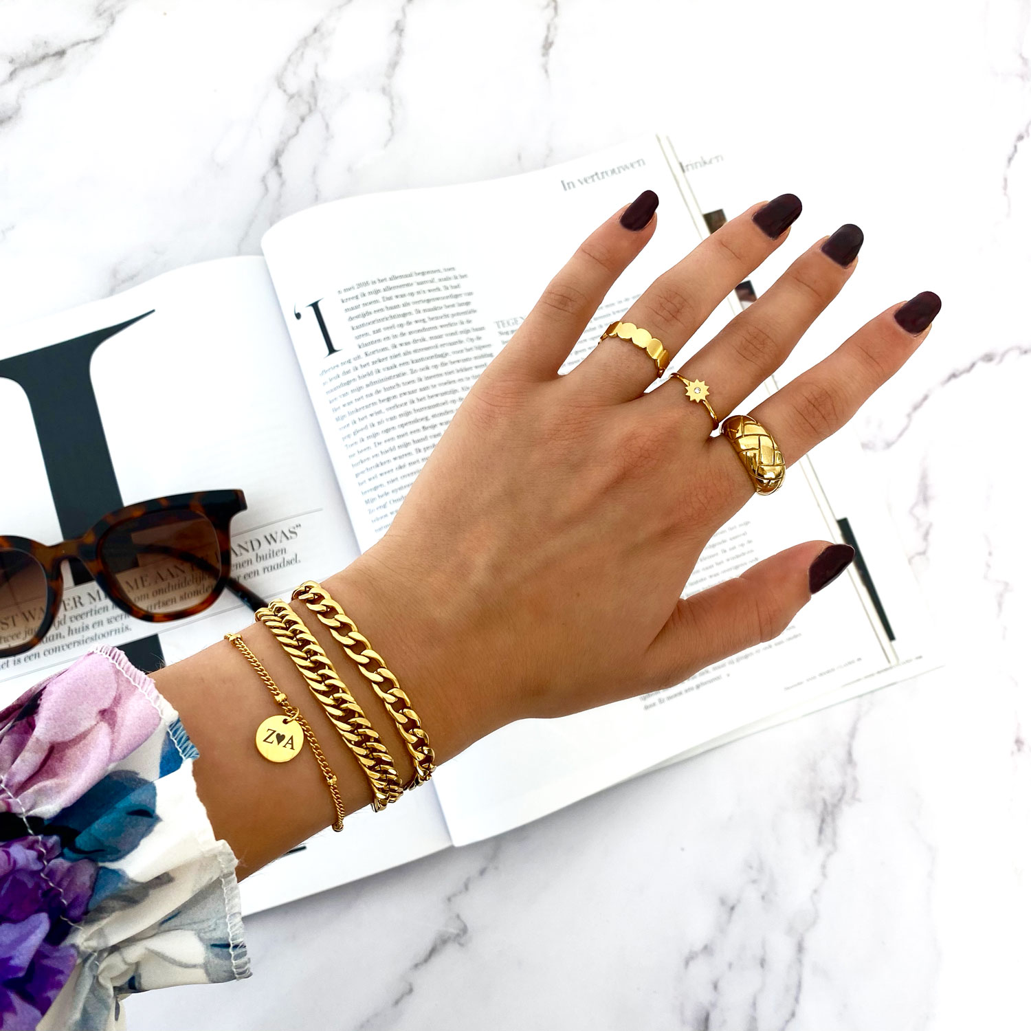 Arm met gouden armbanden set en gouden ringen. Zwarte nagels en een magazine en zonnebril op tafel.